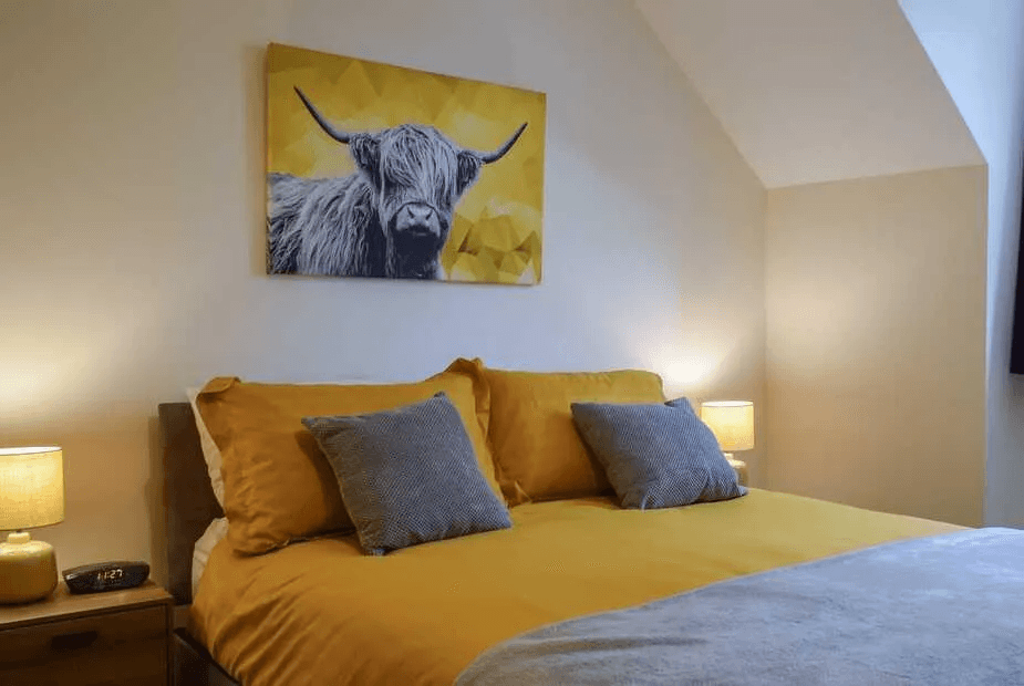 King bedroom at Beechwood with mustard linen, grey cushions and a highland-cow canvas above the headboard