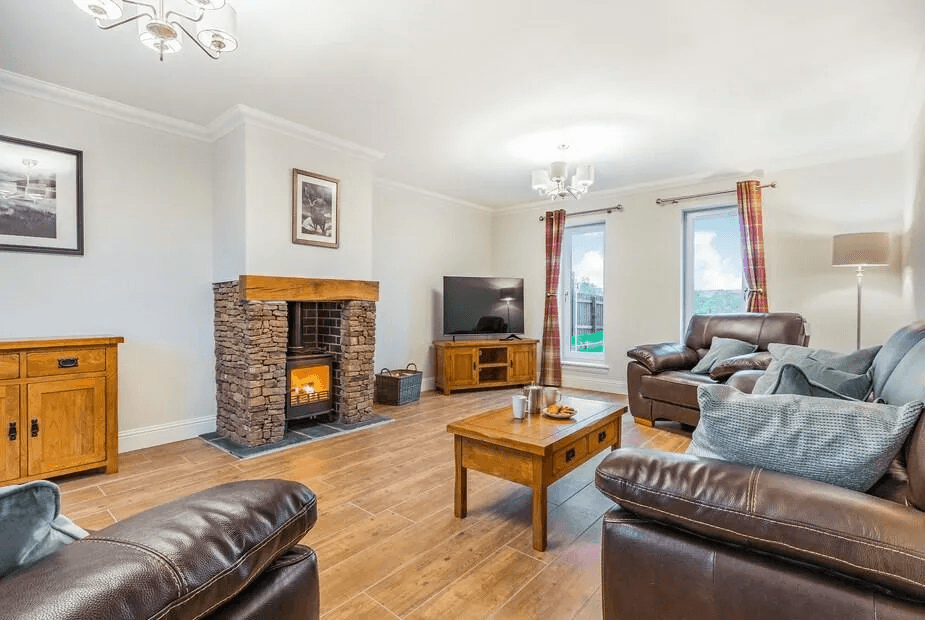 Open lounge at Fiadh Ruadh with two leather sofas, dry-stone fireplace with lit wood-burner and patio doors