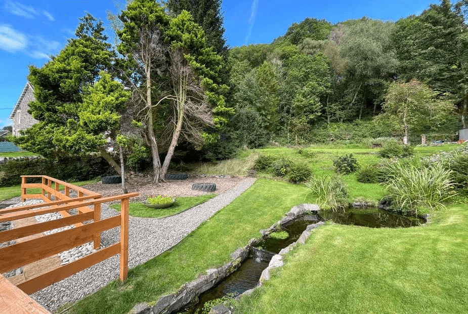 Shared landscaped garden at Holly Apartment with wooden decking, stone-edged pond and stream, and the building visible on the left, Aberfoyle