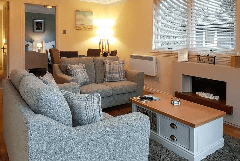 Cosy living room at Leven Lodge 2 with grey sofas, tartan cushions, electric fireplace and bedroom doorway beyond