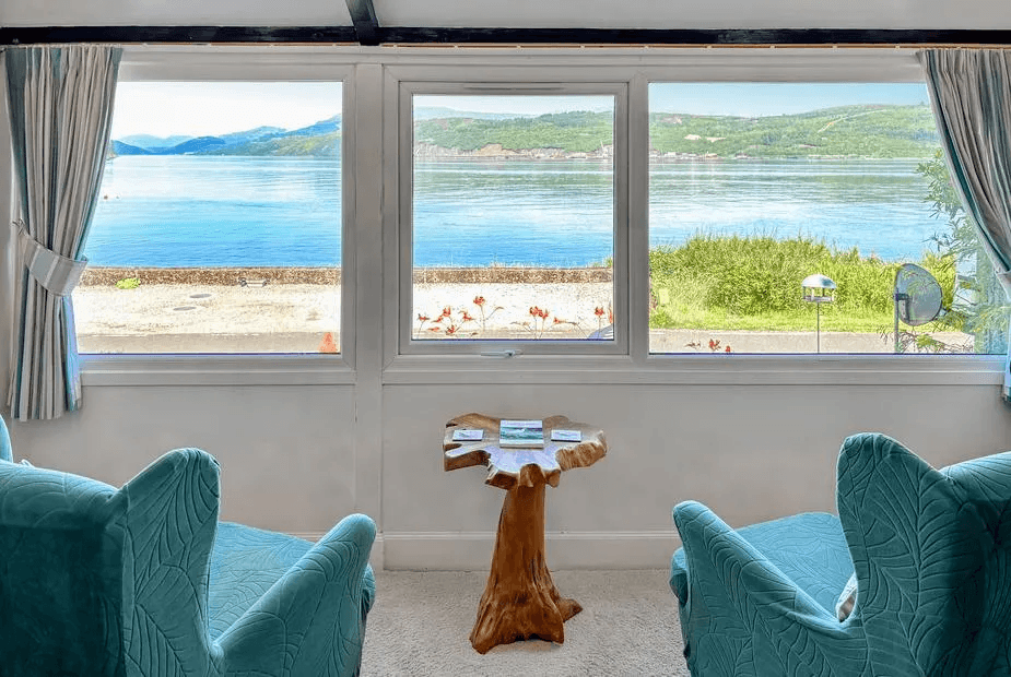 Sitting nook at The Heron with teal velvet armchairs framing a wide loch panorama through corner windows